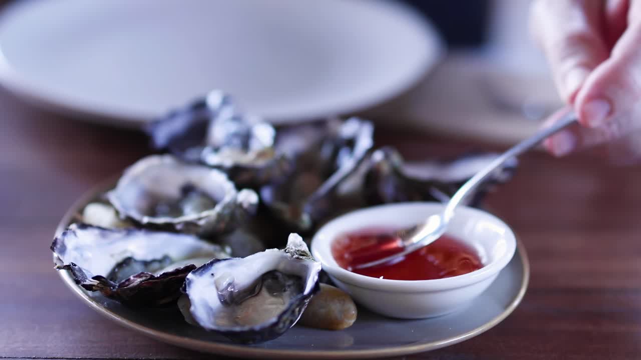 A spoon dips into red sauce beside a plate of fresh oysters on a wooden table.