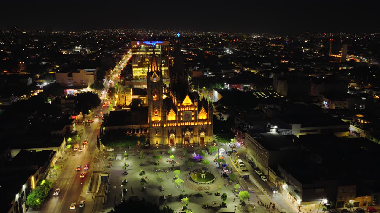 Drone circles Templo Expiatorio at night highlighting intricate Gothic features and colorful stained glass from above, establishing orbit over park in Guadalajara