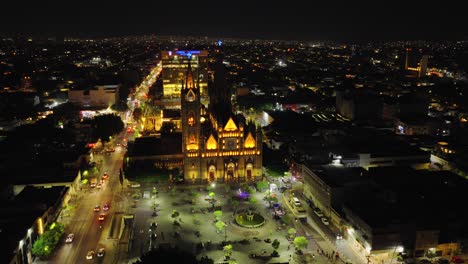 drone circles templo expiatorio at night highlighting intricate gothic features and colorful stained glass from above, establishing orbit over park in guadalajara