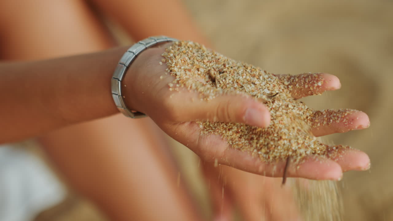 Mano a conca che tiene sabbia calda della spiaggia, luccichio di un braccialetto d'argento, granelli illuminati dal sole che scivolano tra le dita, atmosfera di vacanza rilassata, profondità di campo ridotta con gambe e costa sfocate, esplorazione tattile.