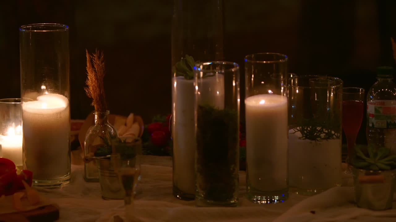 Wedding party banquet outdoors in forest. Dining table decorated in boho style with burning candles, white cloth, flowers, served with plates, tableware, dishes, meals, stemware and drinks at night
