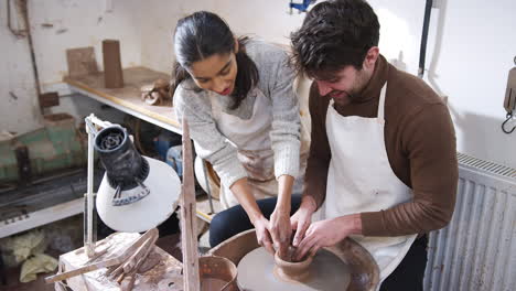 female teacher helping man sitting at wheel in pottery class