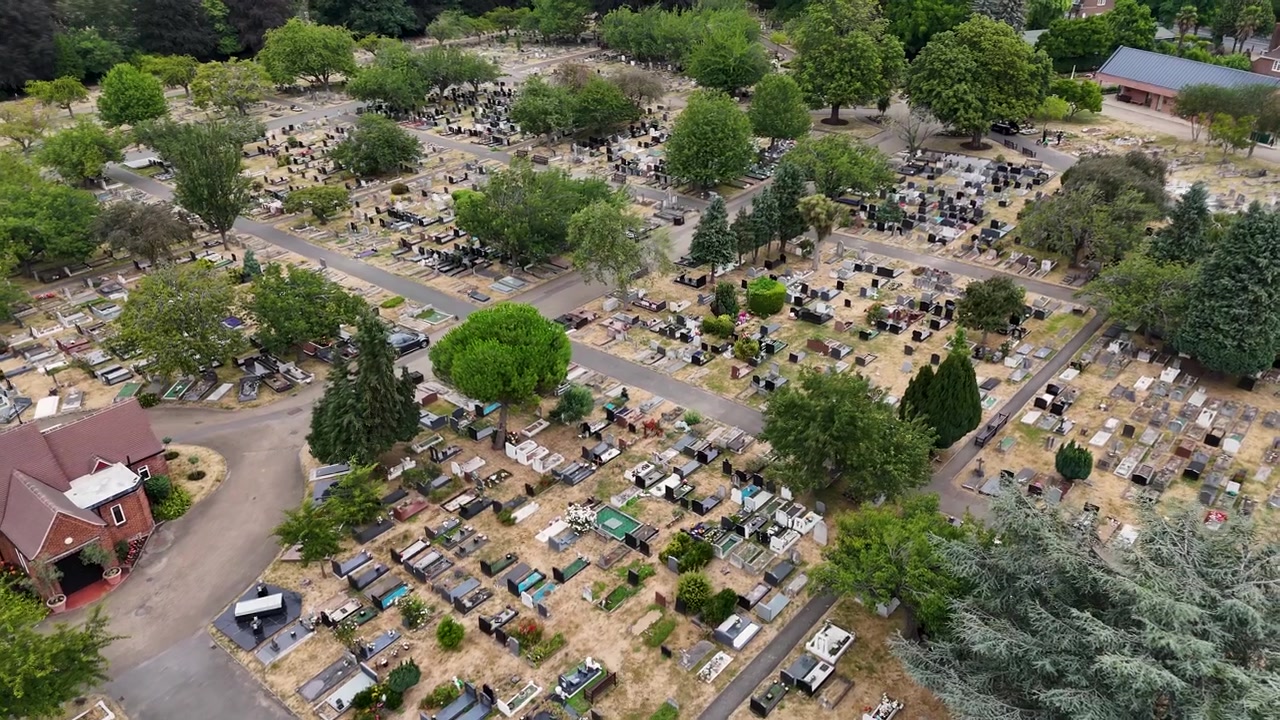 Una piccola cappella si erge tra file di tombe e alberi secolari in un grande cimitero con erba arsa tra le lapidi.