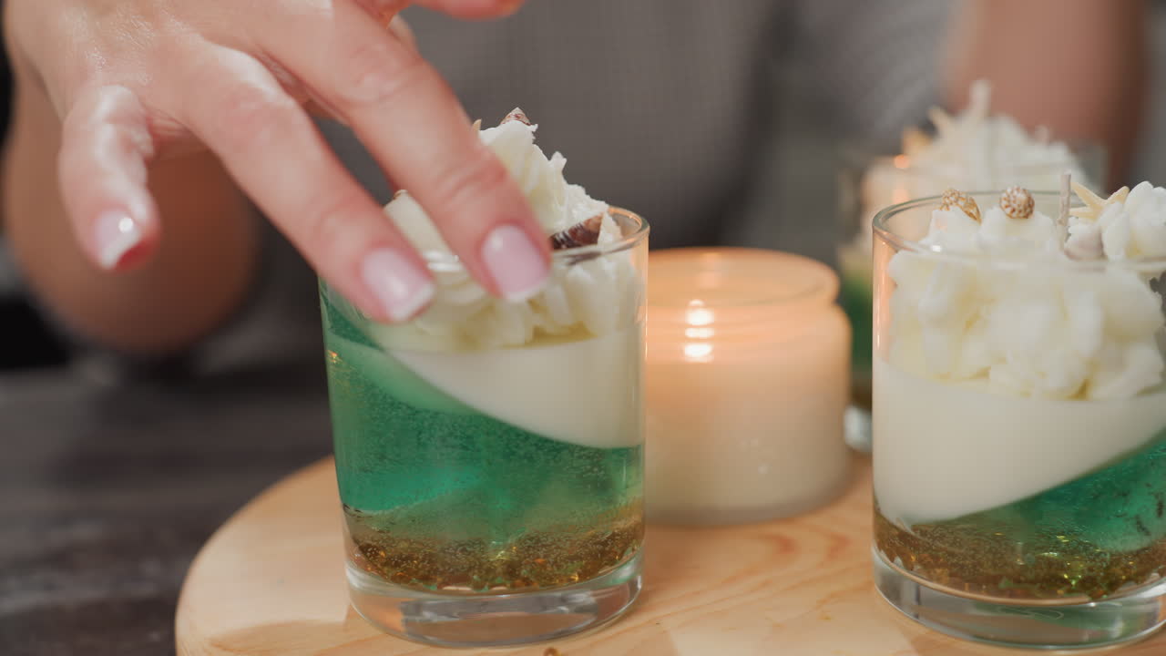 Graceful woman with beautiful manicured hand gently adjusts elegant candle decoration placed on wooden board with sea-inspired elements around, creating cozy peaceful creative atmosphere