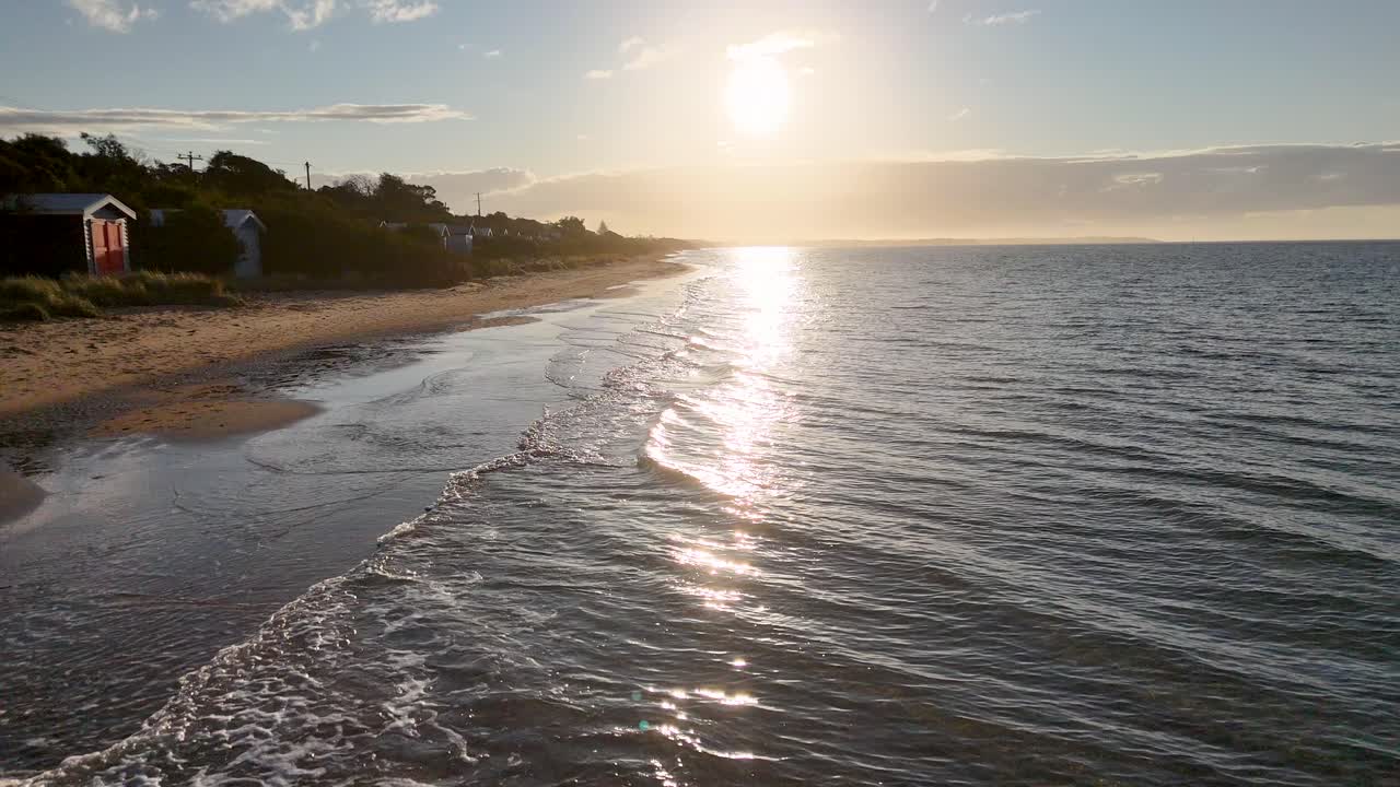 Câmera desliza ao longo da costa ao pôr do sol, passando por cabanas de praia coloridas e ondas suaves do oceano