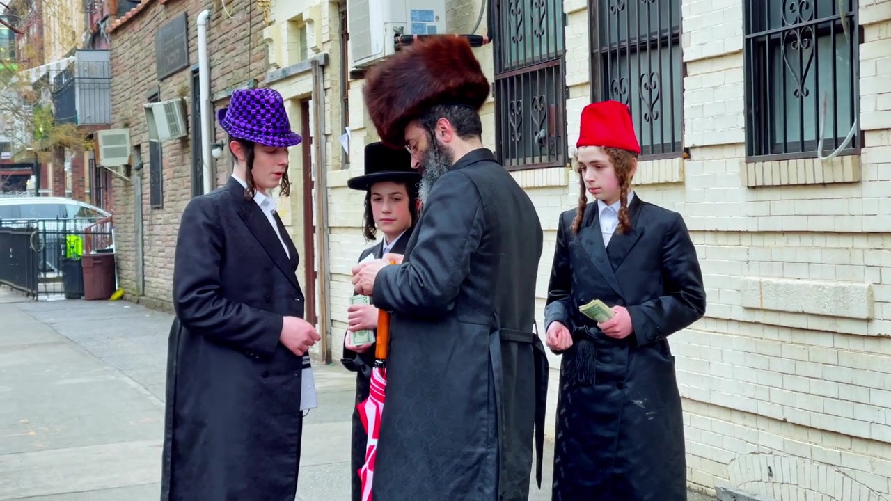 New York, USA, 9 marzo 2022: Gruppo chassidico in piedi sul marciapiede di Brooklyn vicino a edifici in mattoni. Uomini e ragazzi chassidici in cappotti neri stanno sul marciapiede vicino a residenze in mattoni, luce diurna nuvolosa.