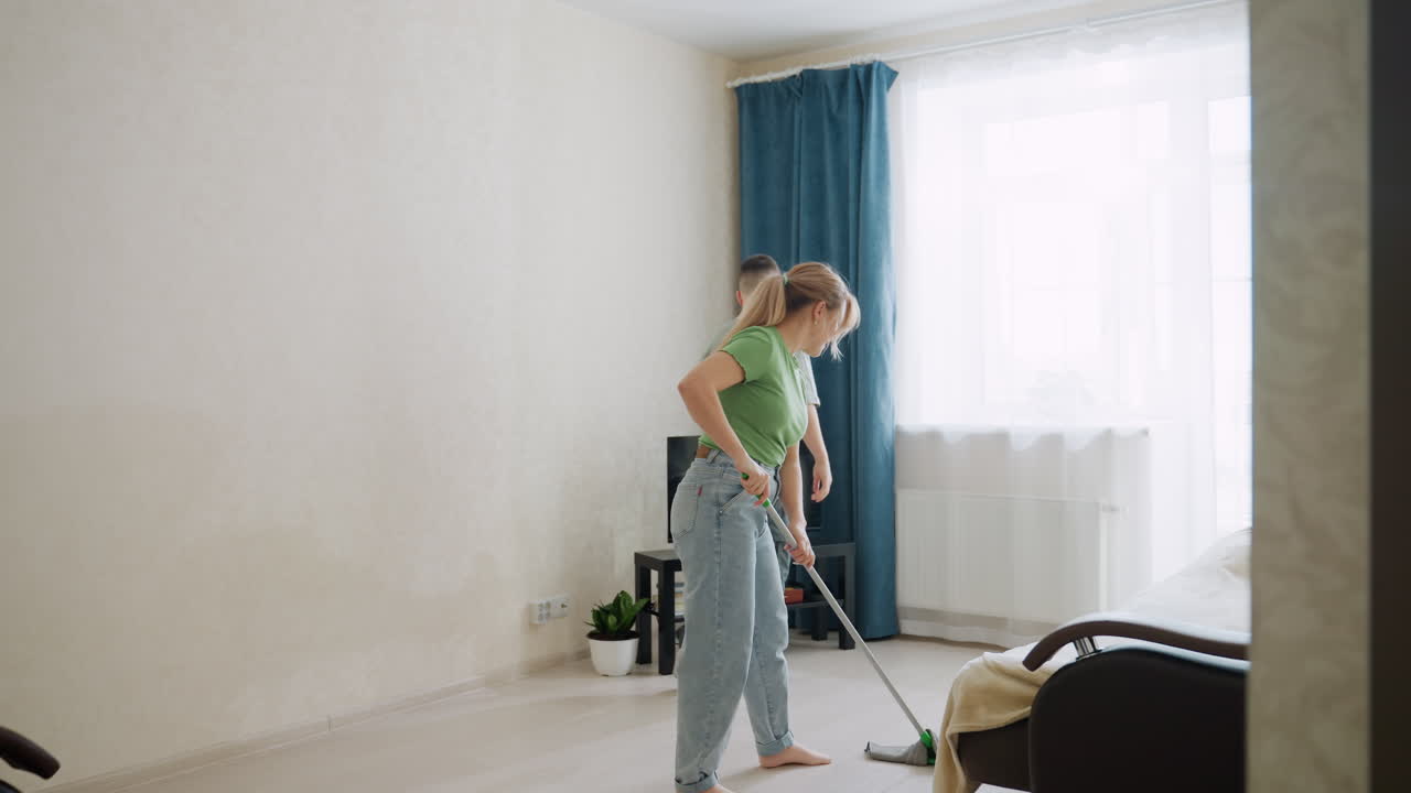 Family tidies up room as woman cleans floor dancing with mop while son dusts television, sunlight through window adds warmth, teamwork turns household chores into joyful bonding activity indoors