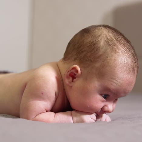 infant lying on the bed on his belly. sweet boy chewing his tiny fists. little kid lying on the bed in a light room close up