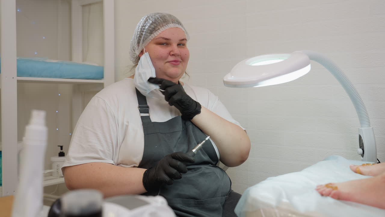 Technician wearing apron and gloves smiles warmly while removing mask and holding electric file under bright salon lamp. She sits beside client feet, exuding confidence and readiness for nail service