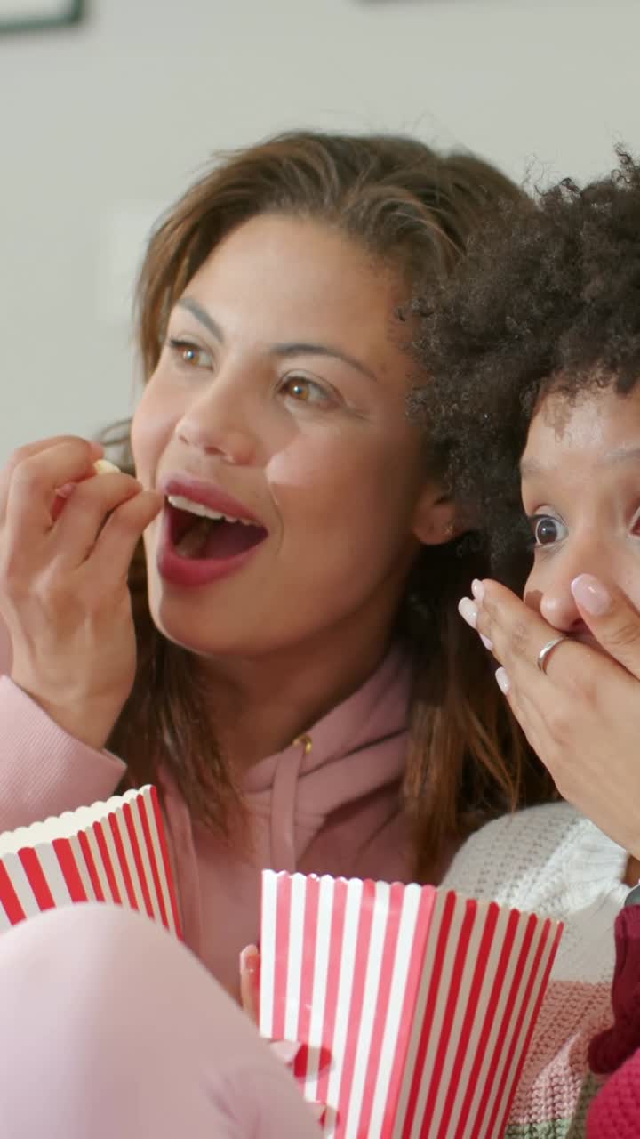 Vertical video: Watching off-screen drama, two women on sofa leaning in, grabbing popcorn, gasping