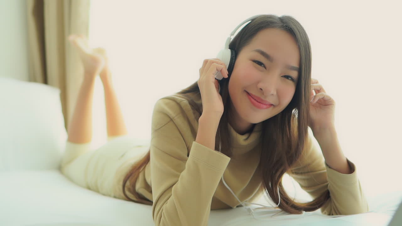 Close -up. Listening to music through her headphone an attractive young Asian woman spontaneously begins to sway and dance to the music while lying on the bed