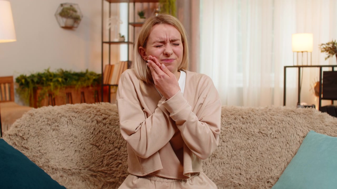 Adult woman sitting on sofa holding cheek with hand showing strong tooth pain discomfort at home