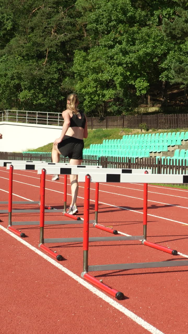 Due giovani donne in forma, in abbigliamento sportivo nero, si allenano su una pista rossa di stadio, saltando gli ostacoli durante una sessione di pratica all'aperto sotto il sole per l'atletica leggera.