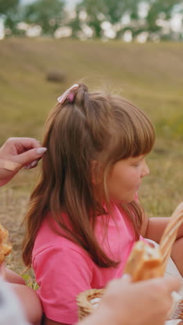 familia pacífica sentada al aire libre disfrutando de bocadillos mientras está rodeada de vastas tierras de cultivo, los niños felizmente comen pasteles de una canasta de picnic mientras comparten un momento relajado en la naturaleza