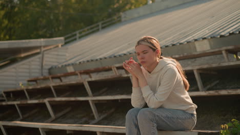 close-up of thoughtful woman in casual hoodie and jeans seated on rustic stadium bleachers with hands joined together, creating a pensive and reflective atmosphere