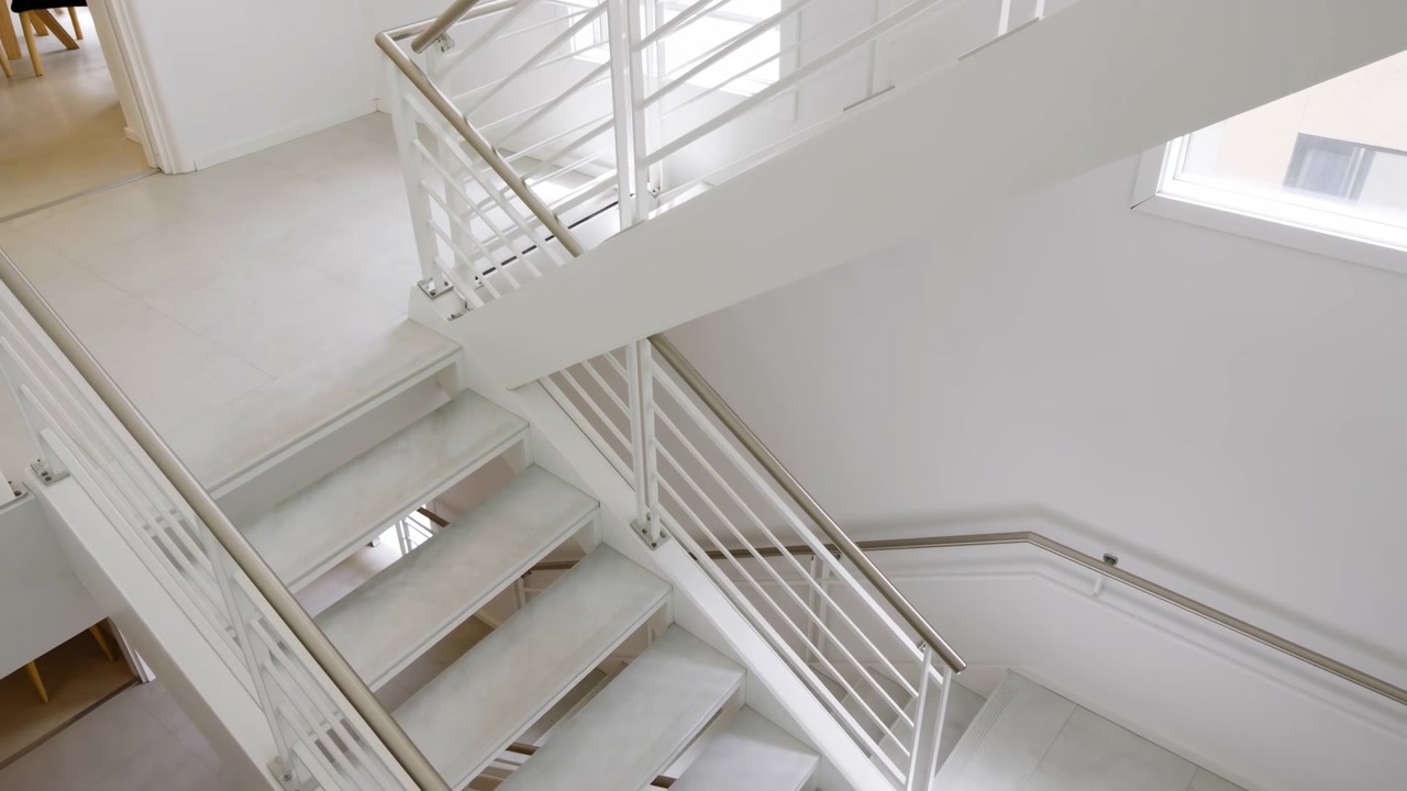 Doğal ışıkla yıkanmış bir iç mekanda, ahşap basamaklar ve metal korkuluk içeren sakin bir merdiven, yukarıdan aşağıya bir perspektiften görünerek sakin ve nötr bir atmosfer yaratıyor.