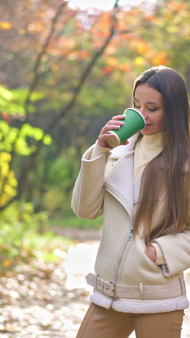 Dark-haired lady in milk-white blazer drinking coffee in the park during walk. Woman taking a stroll alone in the autumn nature.