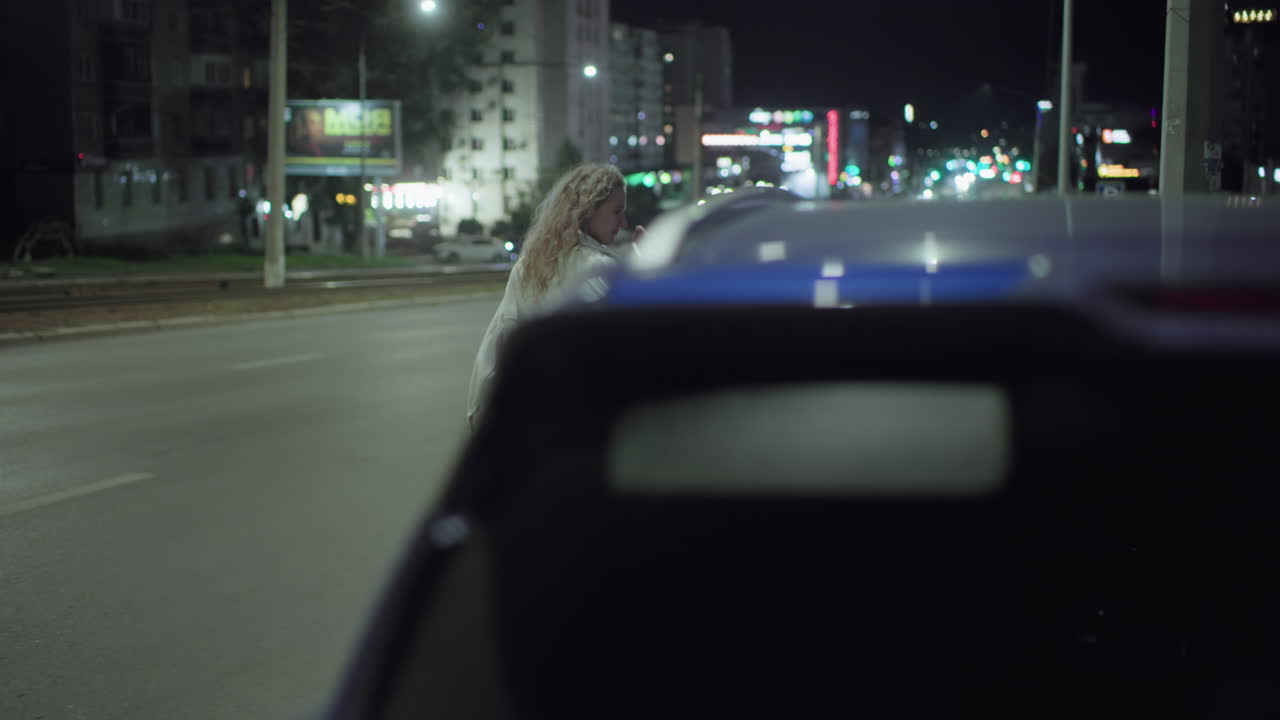 Woman in white coat crosses quiet city street at night, places hand on waist, and walks past parked blue car under streetlight glow with colorful traffic lights and buildings in background