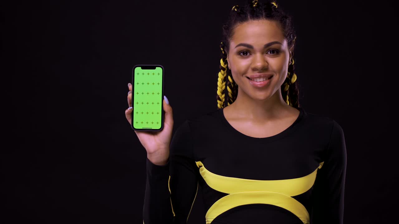 retrato de una chica afroamericana sonriente apuntando con el dedo a la pantalla del teléfono móvil con una llave cromática. mujer aislada sobre un fondo negro
