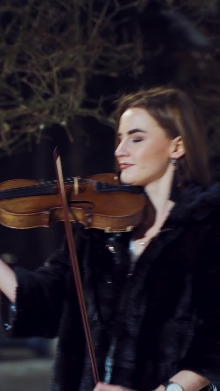 A beautiful girl with make-up and in a white dress is playing a violin composition in a winter park on the background of snow. Camera motion to left Vertical video