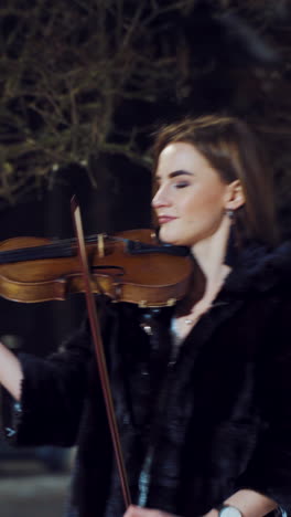 a beautiful girl with make-up and in a white dress is playing a violin composition in a winter park on the background of snow. camera motion to left vertical video