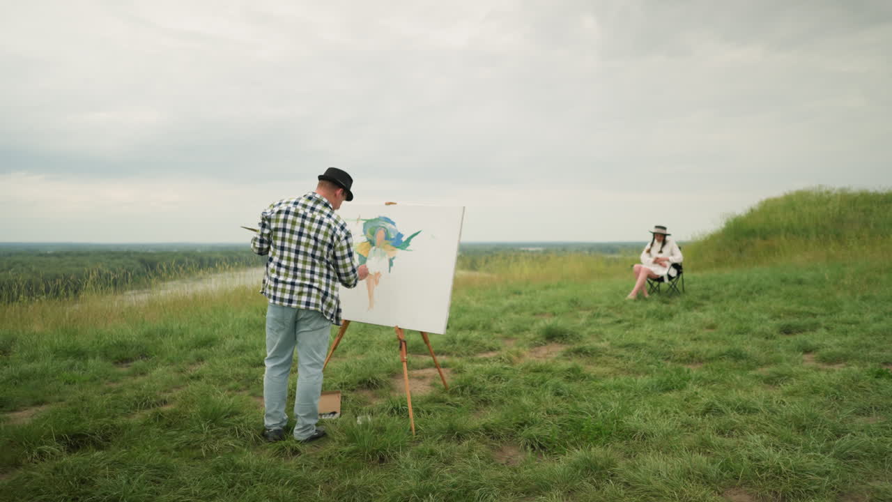 Painter in a hat and checkered shirt, is painting on a board in the middle of a grass field under a cloudy sky. a woman in a hat and white dress sits on a chair, with a peaceful lake in the background