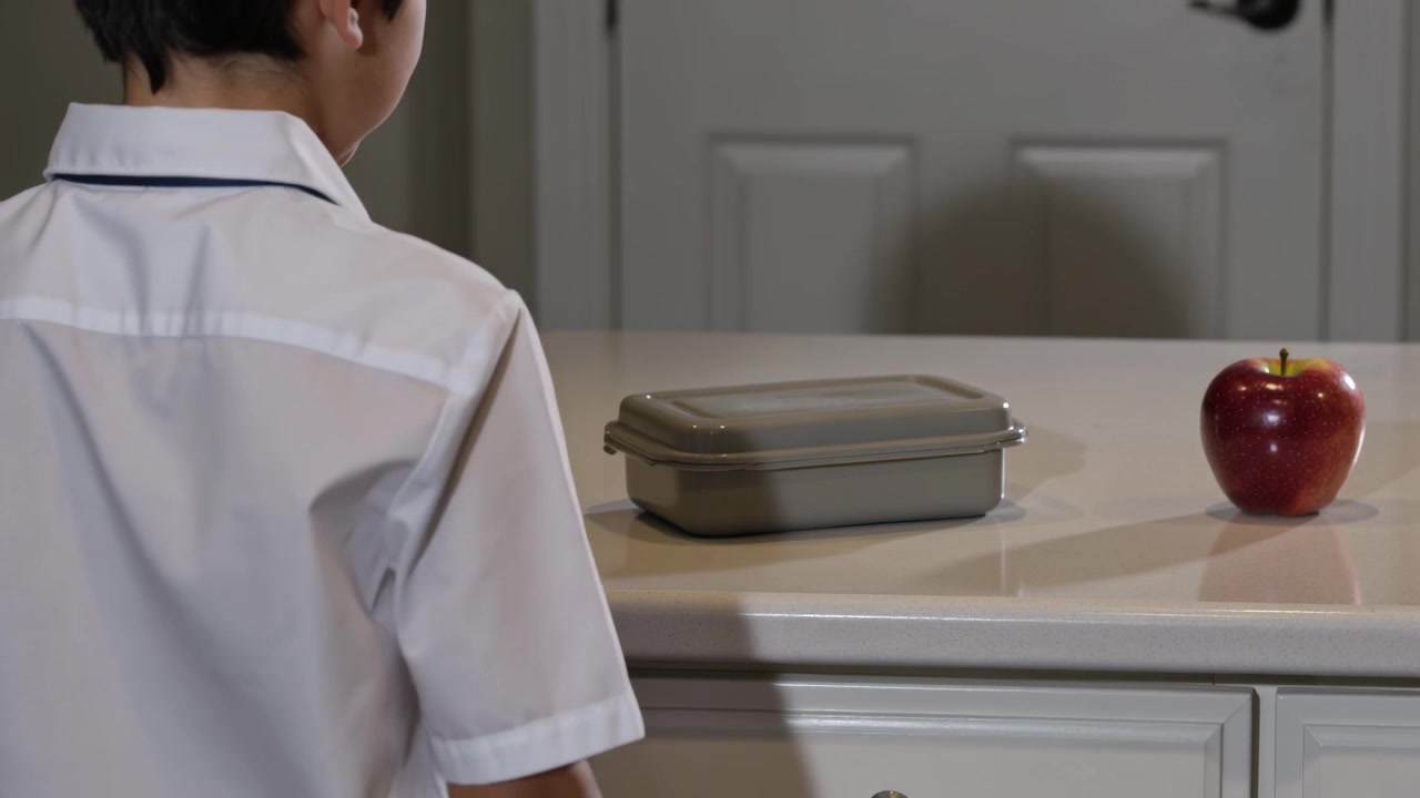 Placing lunch container, kid in white collared shirt grasping red apple at kitchen counter, packing