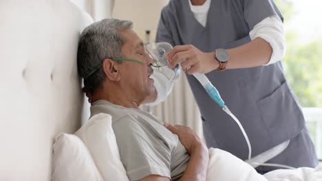 médico varón diverso ayudando a un paciente varón anciano usando una máscara de oxígeno en casa, cámara lenta