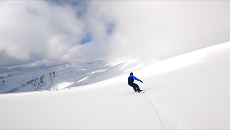 following a male snowboarder riding by the beautiful snowy landscape. thick cloudscape covering the sky above the mountains.