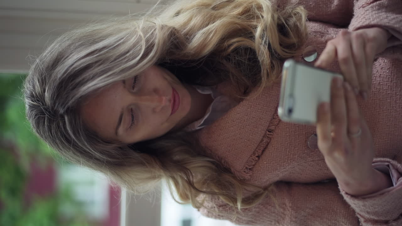 Blonde woman in a pink jacket taking pictures of herself and scrolling through her phone. Vertical