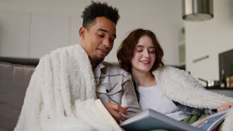 A-happy-young-man-with-Black-skin-color-a-brunette-in-a-checkered-beige-shirt-is-sitting-on-the-sofa-with-his-young-adult-brunette-girlfriend-with-a-bob-hairstyle-wrapped-in-a-white-woolen-blanket.-They-are-reading-a-book-and-relaxing-in-a-modern-apartment