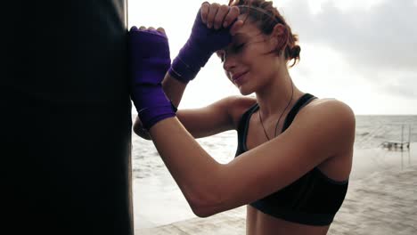 Close-Up-view-of-a-young-woman-having-a-break-after-hard-training-by-the-boxing-bag-against-the-son.-Her-hands-are-wrapped-in