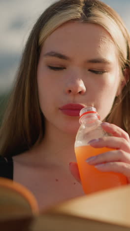 mujer sentada al aire libre, sosteniendo un libro en una mano mientras beba jugo de una botella en la otra, continúa leyendo, con el libro ligeramente borroso y un fondo sereno y borroso