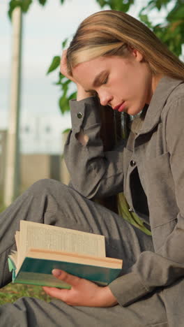 dama sentada al aire libre bajo un árbol, leyendo un libro con la mano debajo del libro, las piernas estiradas, la cabeza ligeramente inclinada con la cabeza apoyada en la mano, la vegetación y la gente borrosa caminando en el fondo