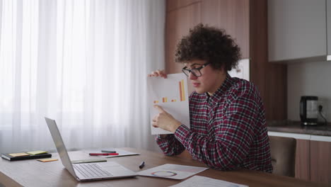 Curly---haired-with-glasses-business-man-sitting-at-office-from-home-desk-looking-at-camera-and-pointing-at-a-tablet-with-financial-information-displayed-in-graphical-form-column-graph