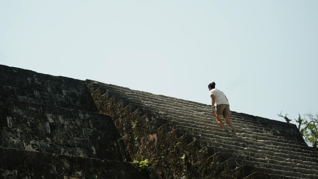 Young man ascends ancient stone steps of a Mayan temple pyramid at Tikal National Park under clear skies.