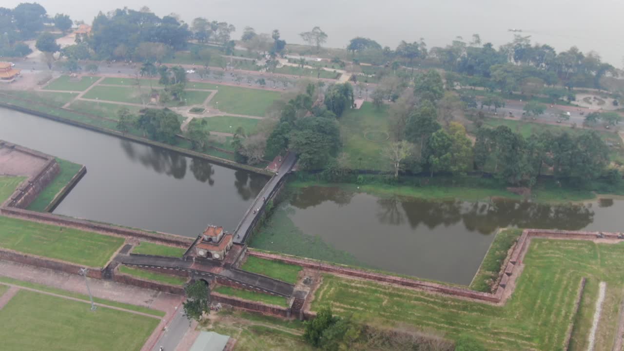 vista aérea de drones en vietnam volando sobre la muralla imperial de ladrillo de piedra, jardines verdes, templos, canales y un amplio río en un día nublado y brumoso