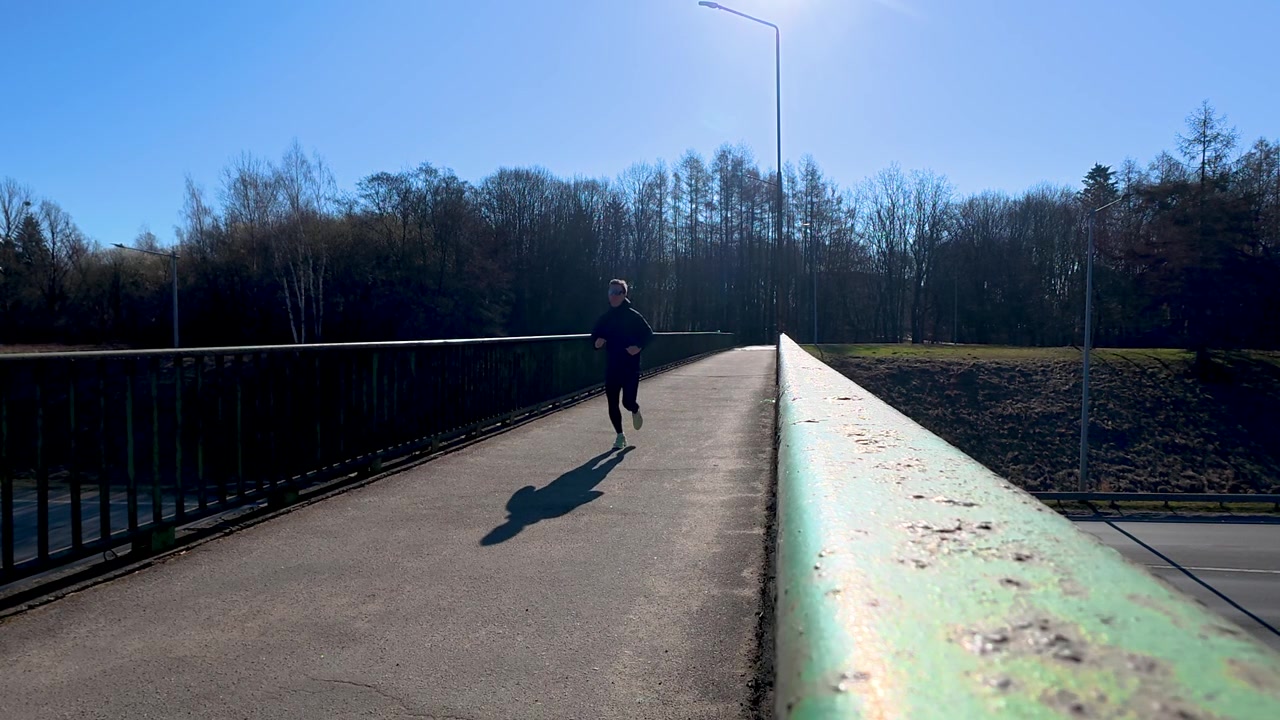 A solitary jogger crosses a bridge under clear blue skies, with bare winter trees in the background. The bright sunlight enhances the tranquility of the scene.