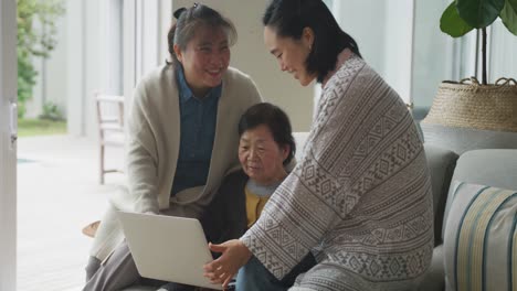 happy senior asian mother, adult daughter and granddaughter sitting in living room using laptop