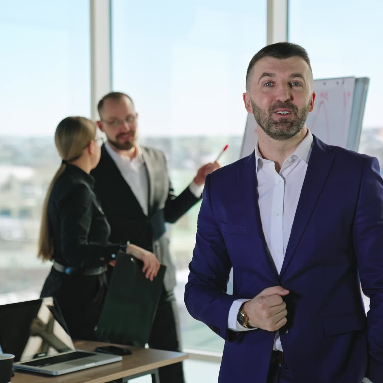 Bearded businessman smiling and talking to the camera. Successful professional standing in the office. Two office people at the backdrop
