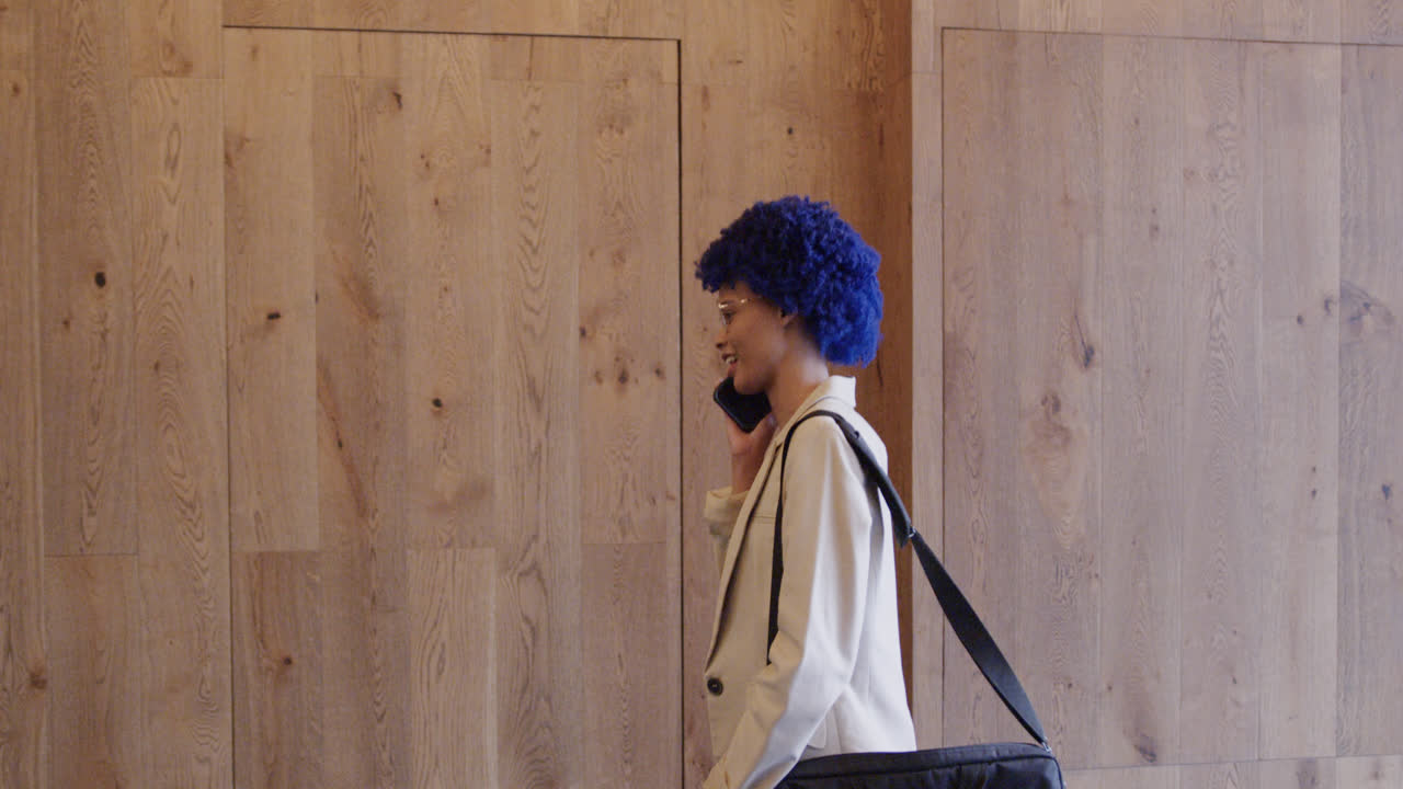 Happy biracial businesswoman with blue afro talking on smartphone in office, slow motion
