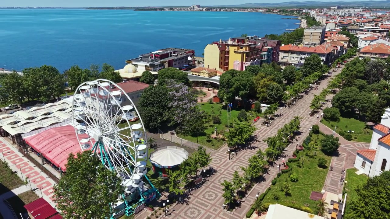 Pessoas desfrutam de um dia ensolarado perto da orla, com uma roda-gigante e uma paisagem urbana ao fundo, capturando a vida local durante a tarde.