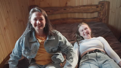excited ladies laugh joyfully while lying on brown blanket inside cozy wooden room, arms spread wide and faces glowing with happiness, sharing spontaneous carefree moment full of laughter