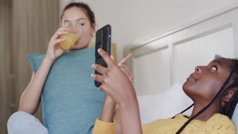 happy diverse teenage female friends lying on bed with drink, talking and using smartphone