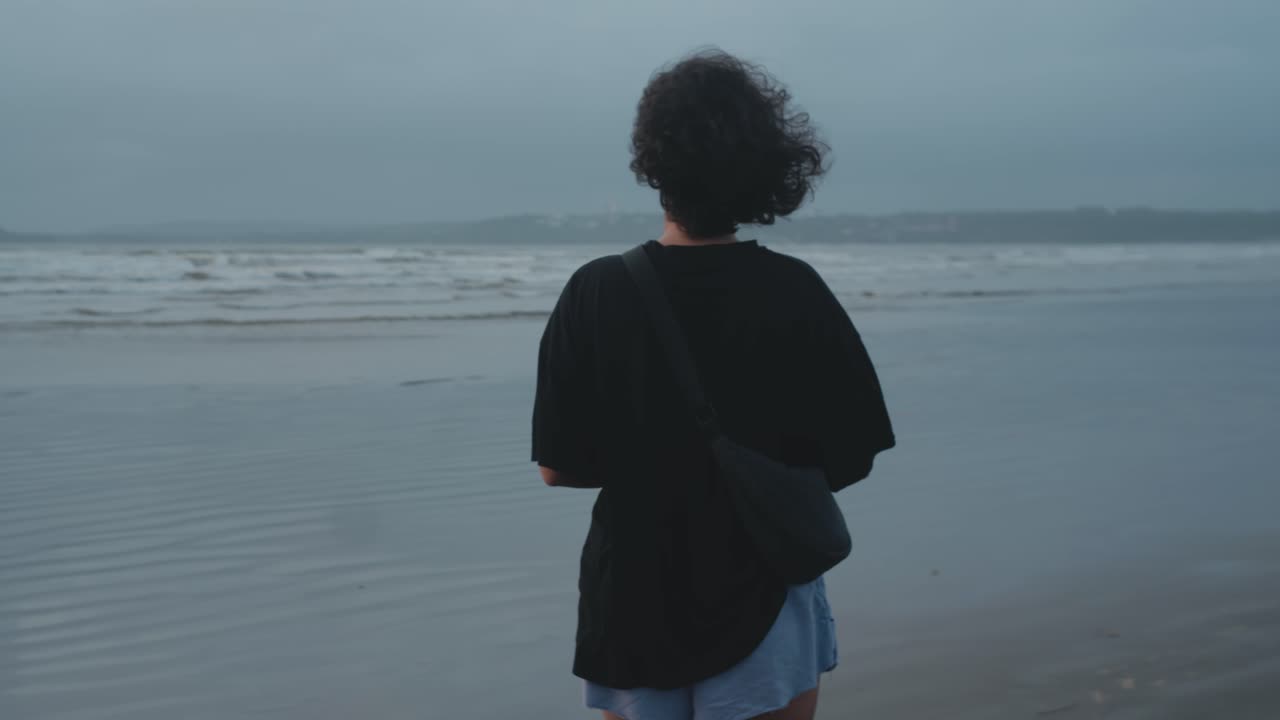 Following a woman walking along a grey windy beach as small waves roll gently across the shoreline