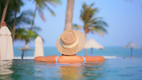 Vista-Trasera-De-La-Joven-En-La-Piscina-Infinita-Con-Un-Sombrero-De-Paja-Y-Apoyando-Los-Brazos-En-El-Borde-De-La-Piscina-En-Un-Hotel-Tropical-En-Malasia