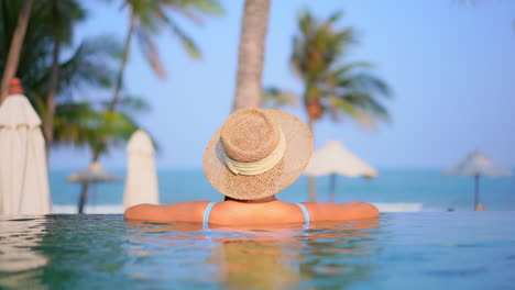 back view of the young woman in infinity swimming pool wearing a straw hat and leaning her arms on the edge of the pool in tropical hotel in malaysia