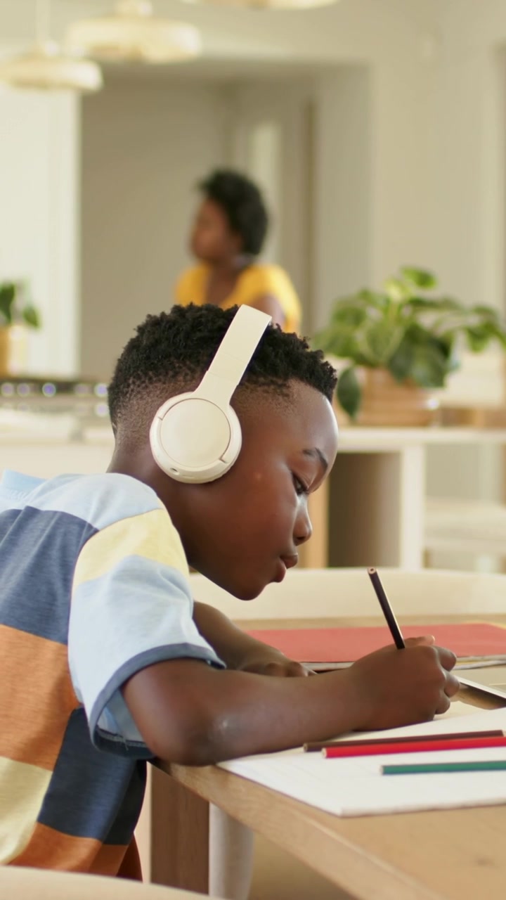Vertical video: Writing boy striped shirt at kitchen table, doing homework with pencil, headphones