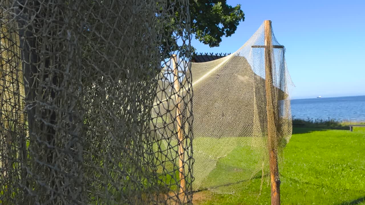 Changing focus footage of gorgeous old traditional fishing nets hanging and drying in a seaside shoreline fishing village. Blue Baltic sea ocean and traditional thached roofed log cabins in the back