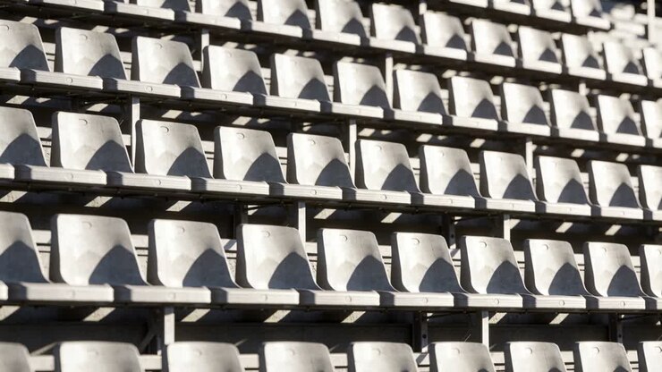 Sunlight shifting, white plastic seats showing changing shadow patterns at grandstand, metal risers